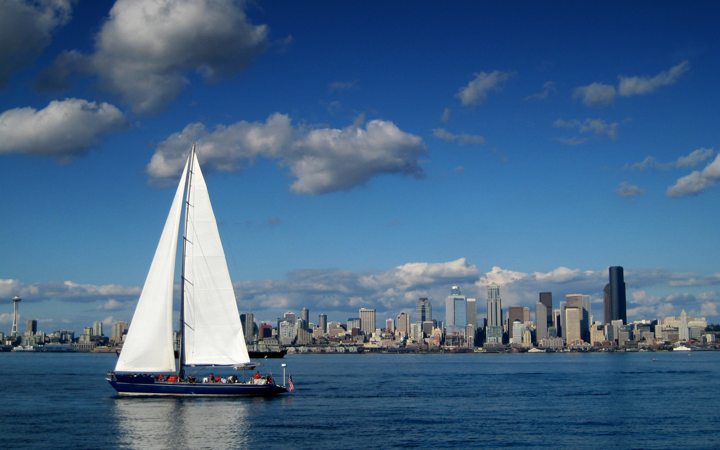 Seattle Skyline with a large sailboat in the foreground with the sails extending above the hoirzon. The Seattle Space Needle frames the left edge of the composition.